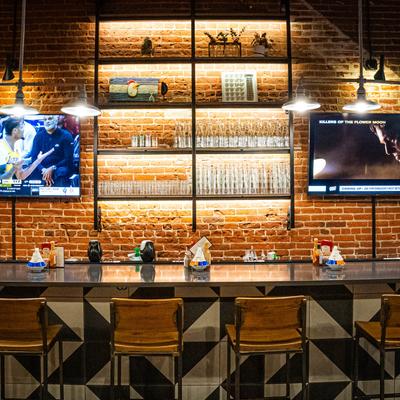 Interior, bar counter, and seats, TV screens and shelves with glasses on a brick wall.