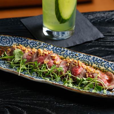 Beef tataki with microgreens on a patterned plate next to a cocktail.
