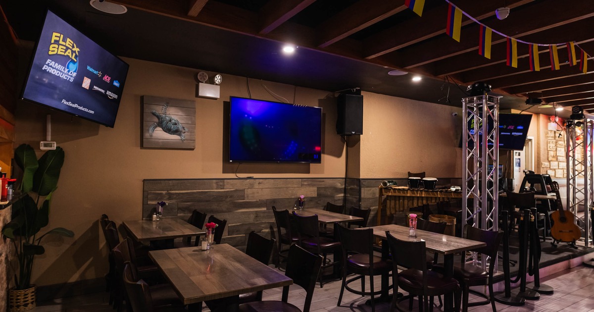 Interior of a restaurant with wooden tables, chairs, TV screens, and a small stage