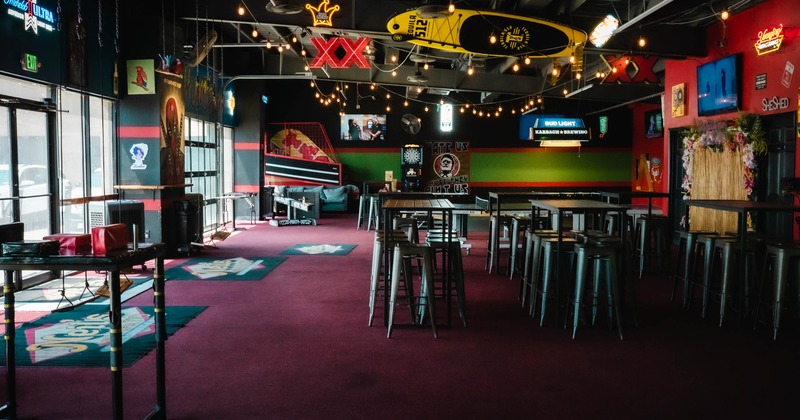 Interior of a modern bar with burgundy carpet, high tables, neon signs, and a suspended yellow surfboard
