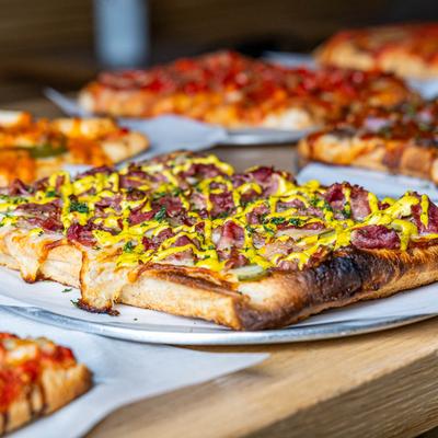 Square-cut pizza topped with bacon, pickles, and mustard drizzle.