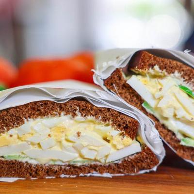 Two halves of an egg sandwich on dark bread, wrapped in paper, are placed on a wooden board.
