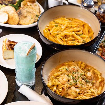 A rich spread of dishes includes pasta, grilled octopus, a cheesecake, and colorful cocktails.