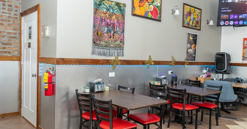Dining area with red-cushioned chairs, dark tables, colorful wall art, blue accent lighting, and a wall-mounted TV