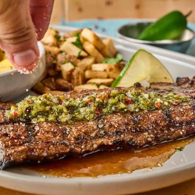 A hand sprinkling salt on a grilled steak topped with chimichurri sauce.