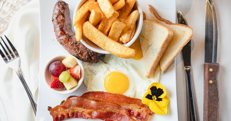 Fried egg, two bacon pieces, sausage, fries and fruit on a breakfast platter