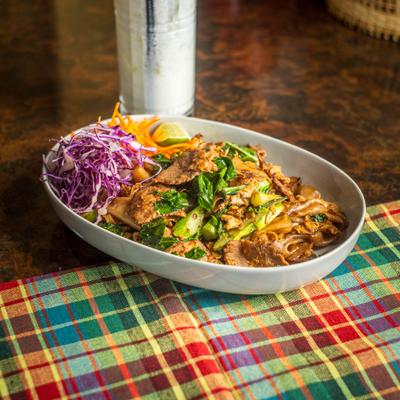 Stir-fried beef noodles with greens and cabbage sit next to a frosted cocktail.