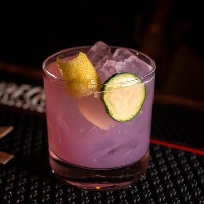 A purple cocktail garnished with a cucumber slice and lemon peel.