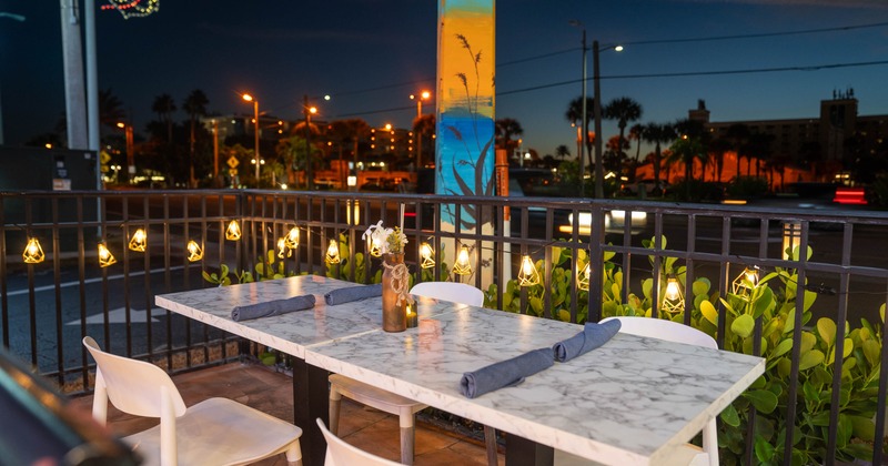 Outdoor dining area with a marble table, white chairs, string lights and street view at night.