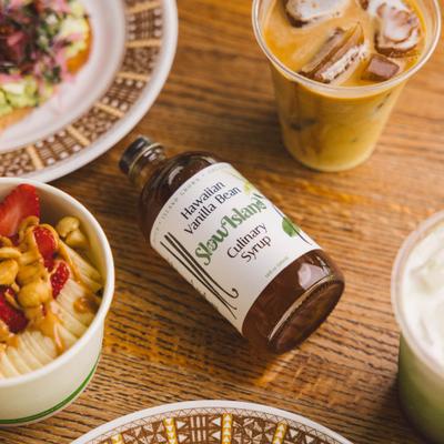 A bottle of Slow Island the Hawaiian Vanilla Bean Culinary Syrup with assorted dishes and beverages.
