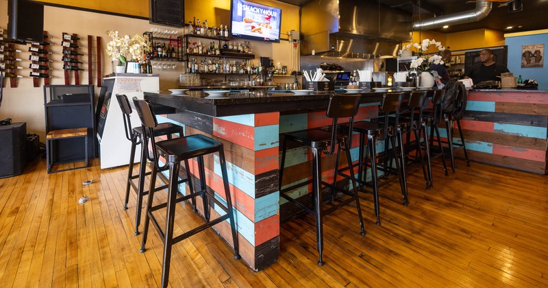 A rustic bar interior with colorful counter, black bar stools, and a wall-mounted TV