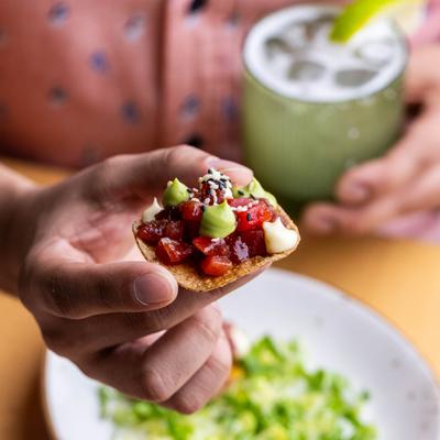 A hand holding a small tostada topped with diced tuna and avocado cream.