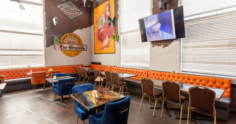 Interior of a restaurant with orange tufted benches, blue velvet chairs, brick walls, retro posters, and a flat-screen TV