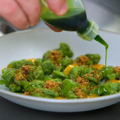 Green gnocchi with brown garnish being drizzled with green sauce in a white bowl.
