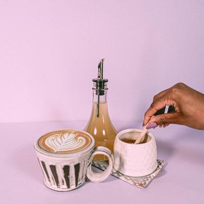 A hand stirring tea beside a latte with art and a syrup bottle.
