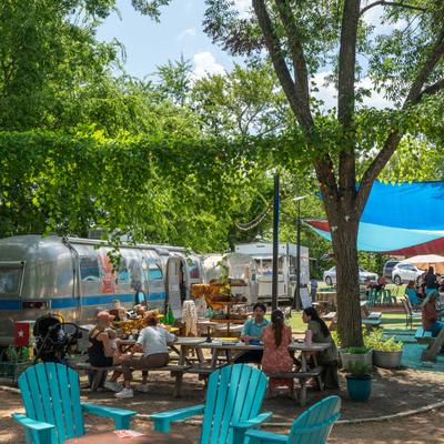 People dining at picnic tables under trees in a food truck park.