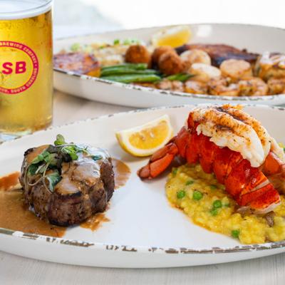 A surf and turf meal featuring steak, lobster tail, risotto, and beer.