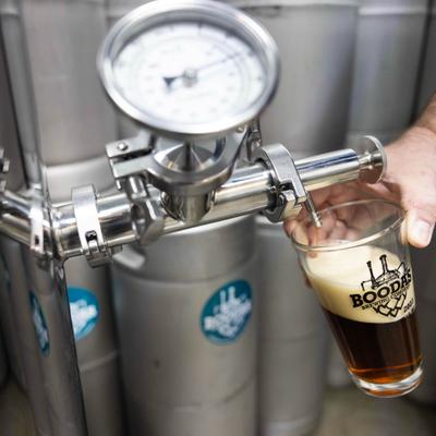 A person pouring a dark beer from a keg tap into a pint glass with the Boodas Brewing logo.