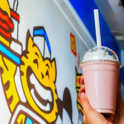 Hands holding a cup with Strawberry Milkshake in fromt of the wall mural.