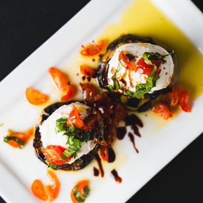 Two grilled eggplant rounds topped with burrata, cherry tomatoes, basil, and balsamic drizzle.