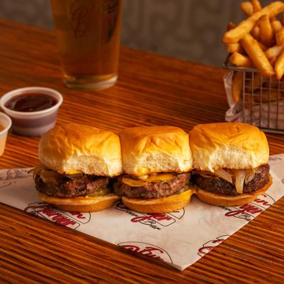 Three beef sliders with cheese and onions served with sauces, fries, and beer.