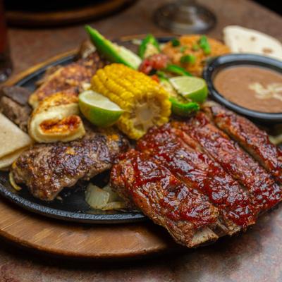 A platter of grilled meats, corn, lime, grilled vegetables, tortillas, and refried beans.