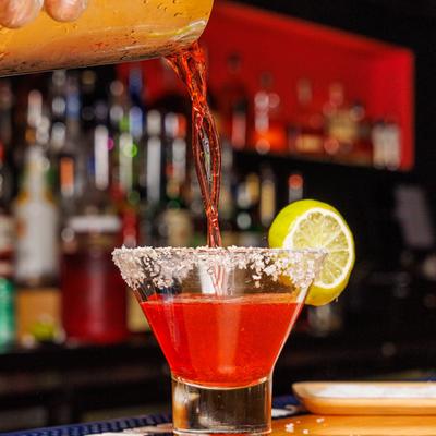 Red cocktail being poured into a stemless martini glass with salted rim and lime garnish.