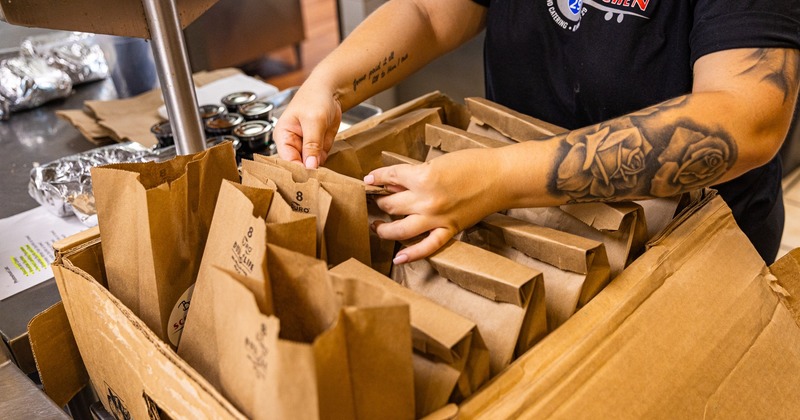 An employee sorts brown paper bags in a cardboard box on a stainless steel countertop