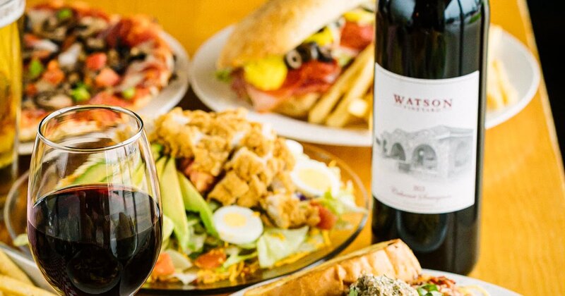 Several dishes of food including pizza, spaghetti and sauce, and a sandwich.  A glass and bottle of wine are included.