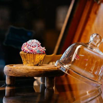 A cupcake with sprinkles on a wooden pedestal next to a dessert glass bell.