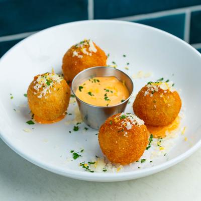 Four golden-brown fried balls & a small cup of orange dipping sauce, garnished with herbs..
