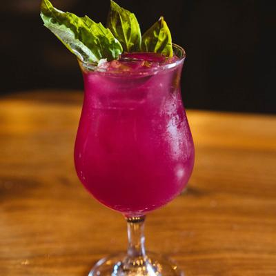 A vibrant pink cocktail, garnished with fresh green basil leaves, sits on a wooden table.