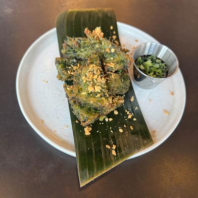 Crispy green fritters on a banana leaf, served with a sauce on a white plate.