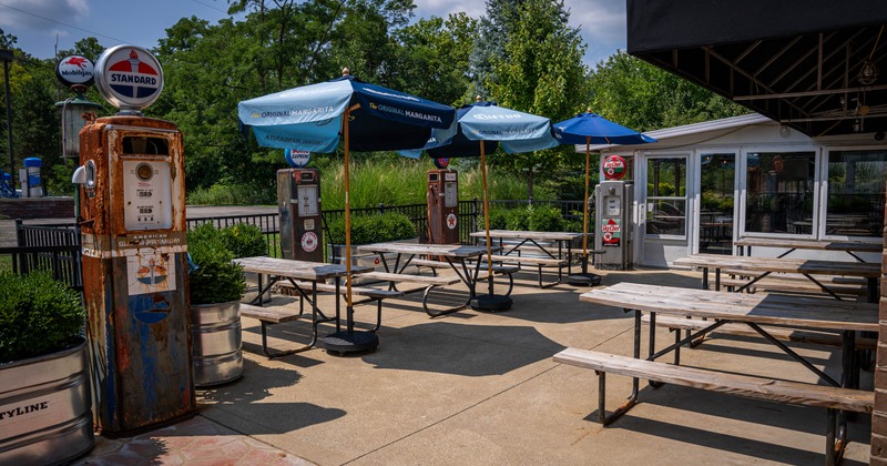 Outdoor dining area with wooden picnic tables, vintage gas pumps, and blue umbrellas
