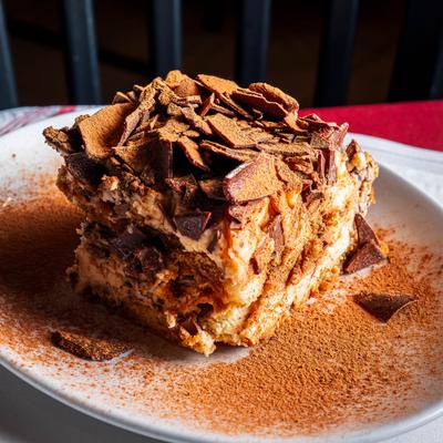 A rich tiramisu slice dusted with cocoa and chocolate shavings sits on a white plate.