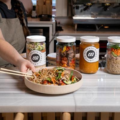 A waitperson serves a plate of duck soba salad on a counter with food jars.