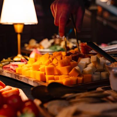 A warmly lit table with a cheese board, a hand uses tongs to serve.