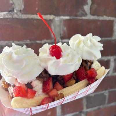 Banana split with whipped cream and cherry on top, held up against a brick wall background.