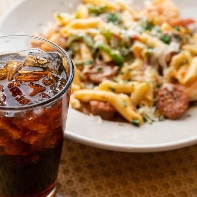 Glass of soda with ice in the foreground and a plate of penne pasta in the background.