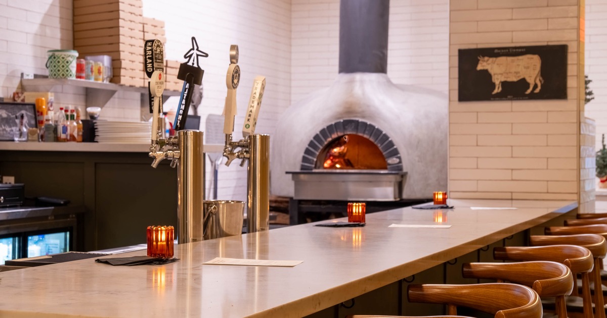 A bar with a stone countertop, wooden bar stools, beer taps and a pizza oven in the background