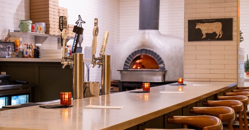 A bar with a stone countertop, wooden bar stools, beer taps and a pizza oven in the background