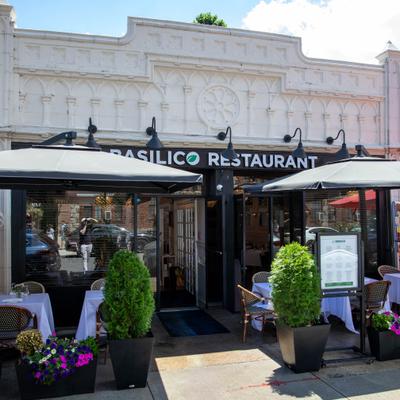Restaurant storefront with outdoor seating, black umbrellas, and potted plants.