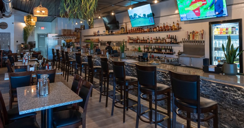 Interior of a modern bar with a long counter, stools, TV screens, and tables