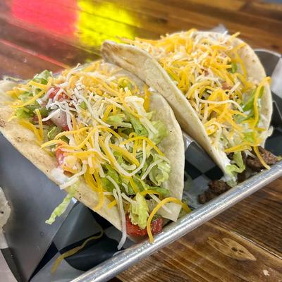 Two tacos filled with seasoned meat, shredded lettuce, diced tomatoes, and cheese sit on a tray.