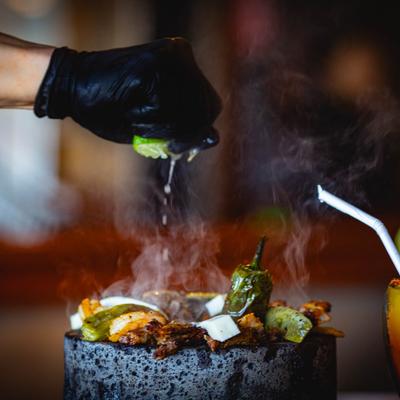 A gloved hand squeezes lime over a steaming molcajete dish.