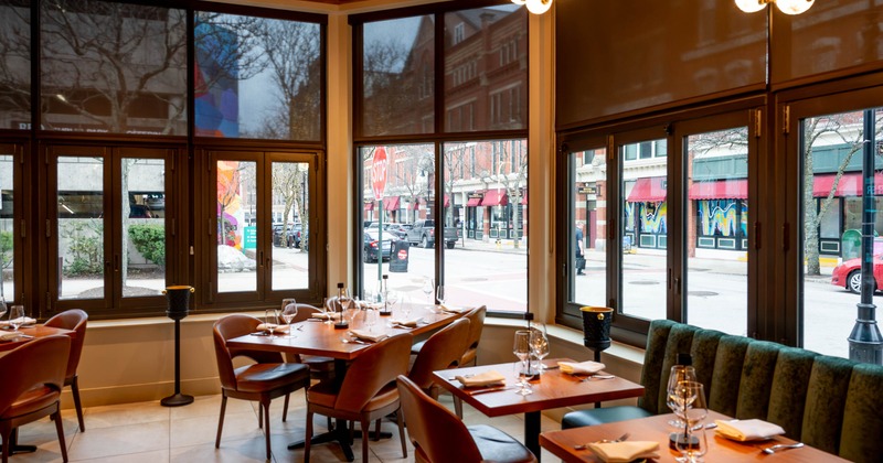 Interior of a restaurant with large windows and set dining tables, overlooking a street with buildings and a stop sign