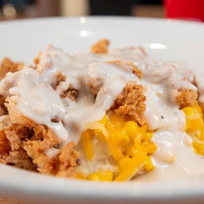 A bowl of mashed potatoes, topped  with corn, gravy, cheddar and fried chicken.