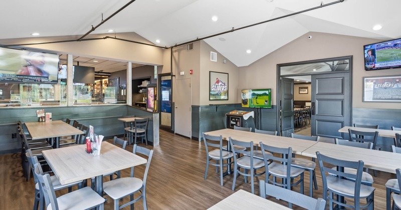 Indoor dining area of a casual restaurant with tables and chairs, two mounted TVs showing sports, and an arcade game