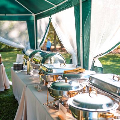 A buffet table elegantly set with chafing dishes and draped in white cloths in a tent.
