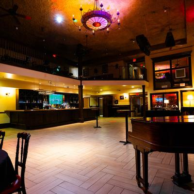 Interior, spacious speakeasy room with seating, a piano, and a bar.